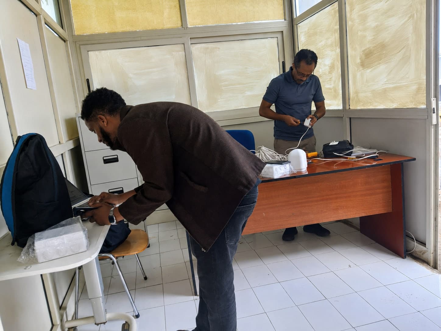 Two engineers indoors preparing a sensor unit for deployment, one working at a laptop while the other connects the enclosure's power lead.