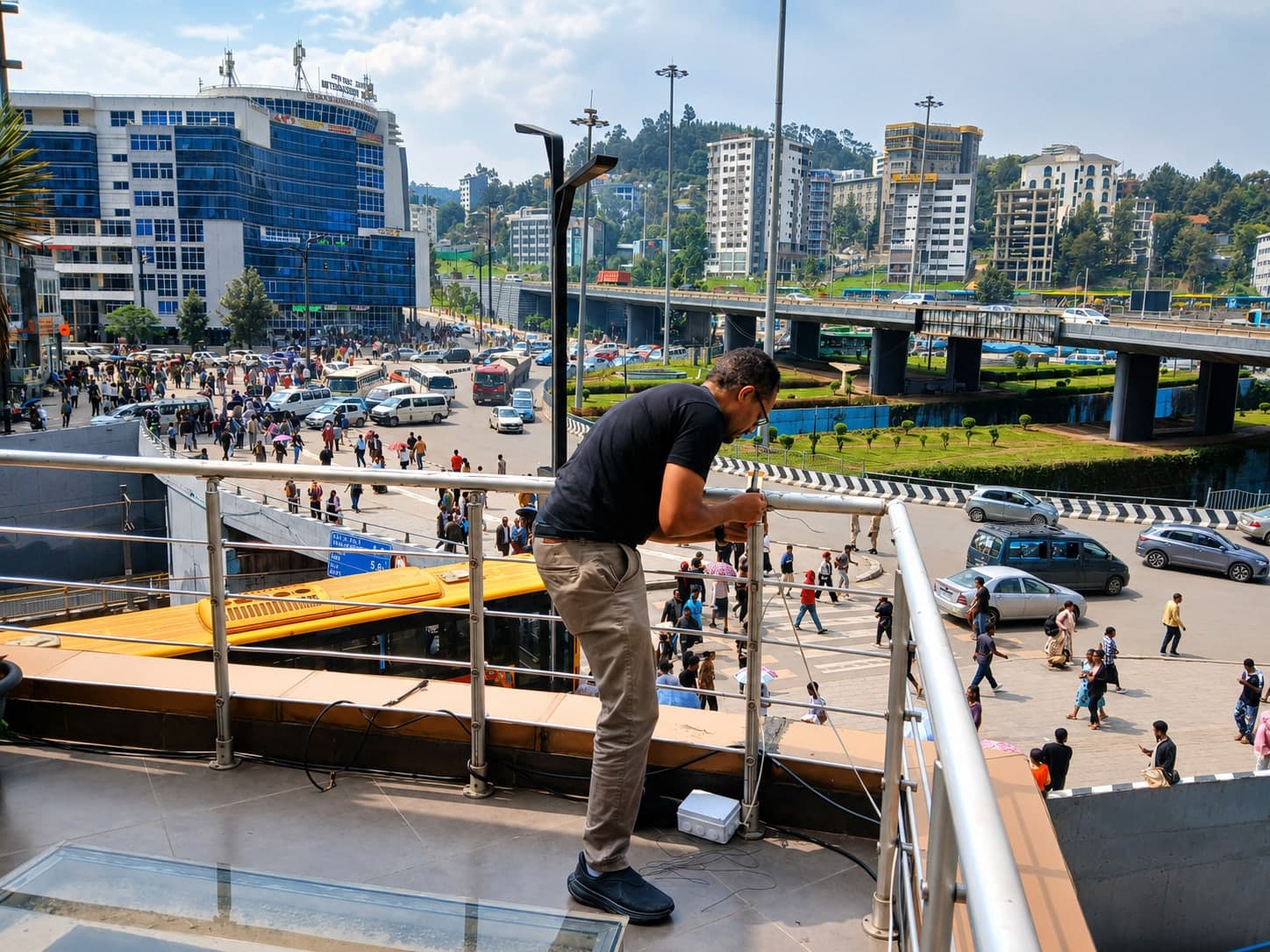 An engineer fastening a sensor mount to the railing of a raised walkway above a busy Addis Ababa intersection, with a bus rapid transit lane, an elevated roadway and high-rise buildings behind.
