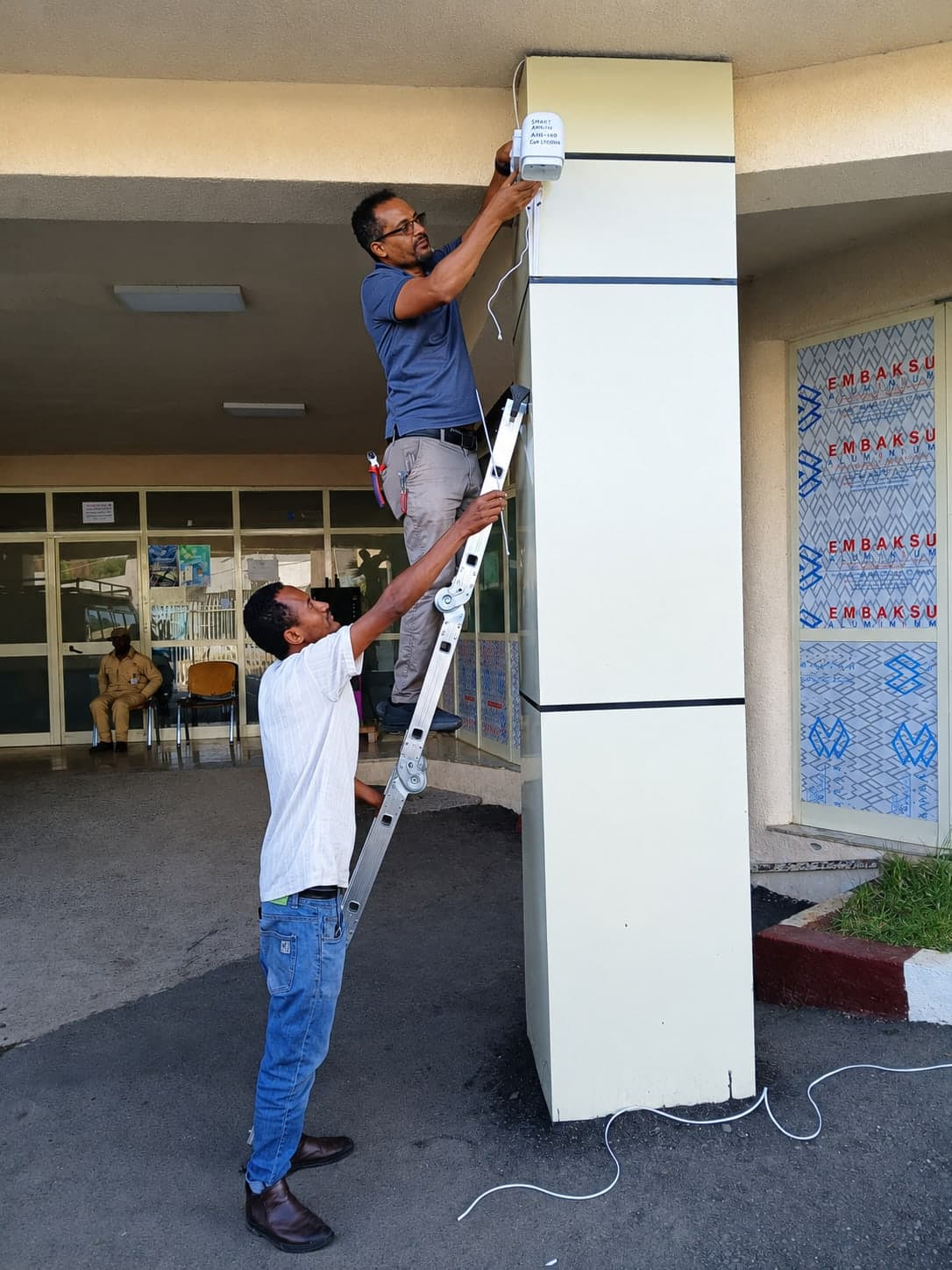 An engineer on a ladder attaching a sensor enclosure to a concrete column at a building entrance while a colleague steadies the ladder.