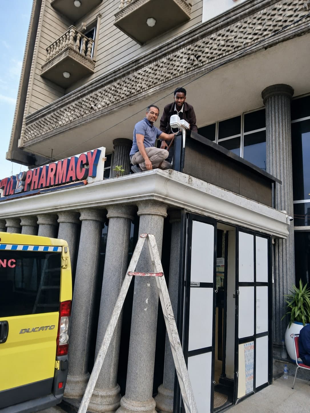 Two engineers on the entrance canopy of a pharmacy building fitting a sensor enclosure above the street.