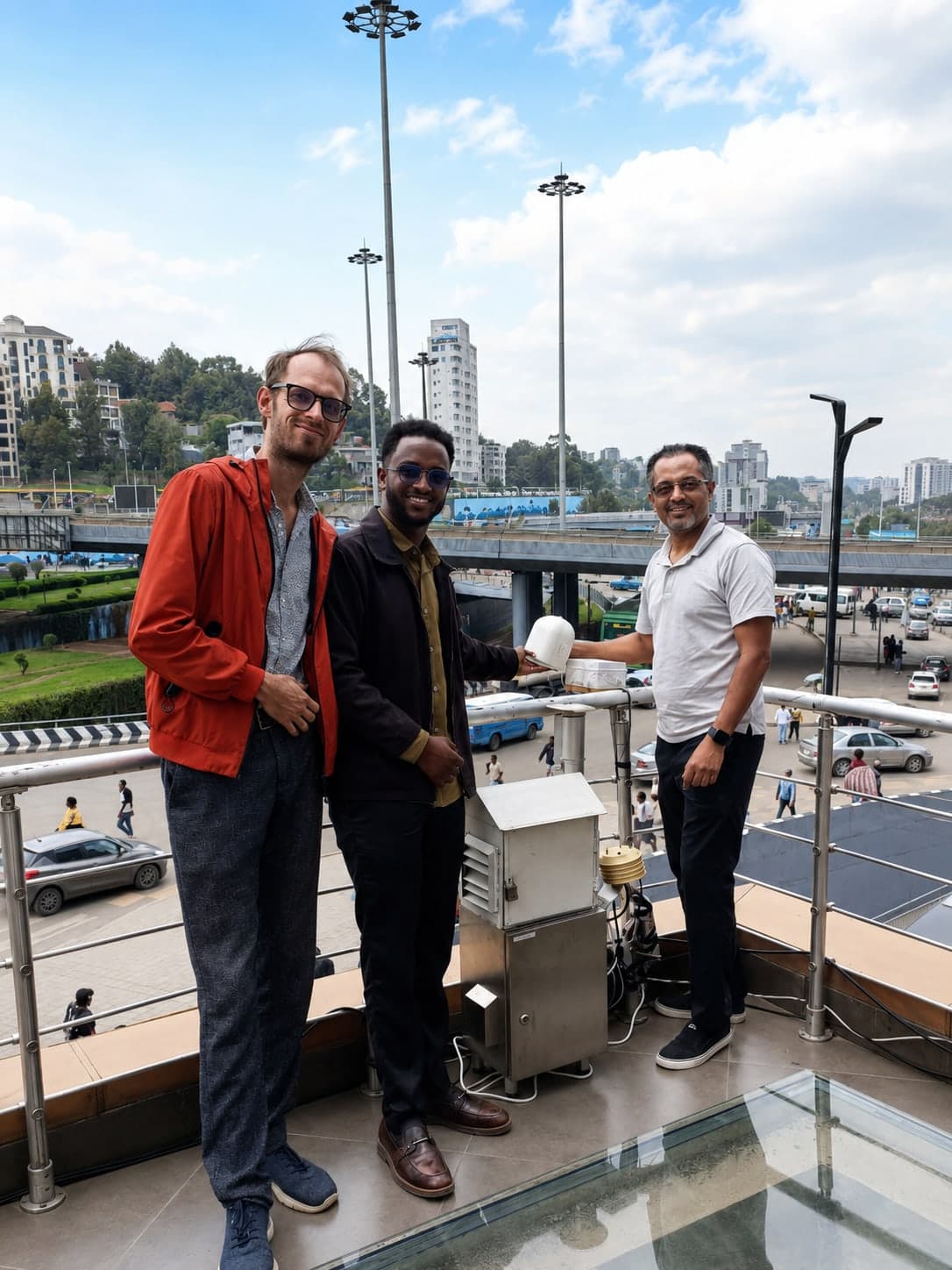 Three members of the project team standing beside a newly installed air quality sensor and its stainless steel equipment cabinet on a walkway overlooking a major interchange.