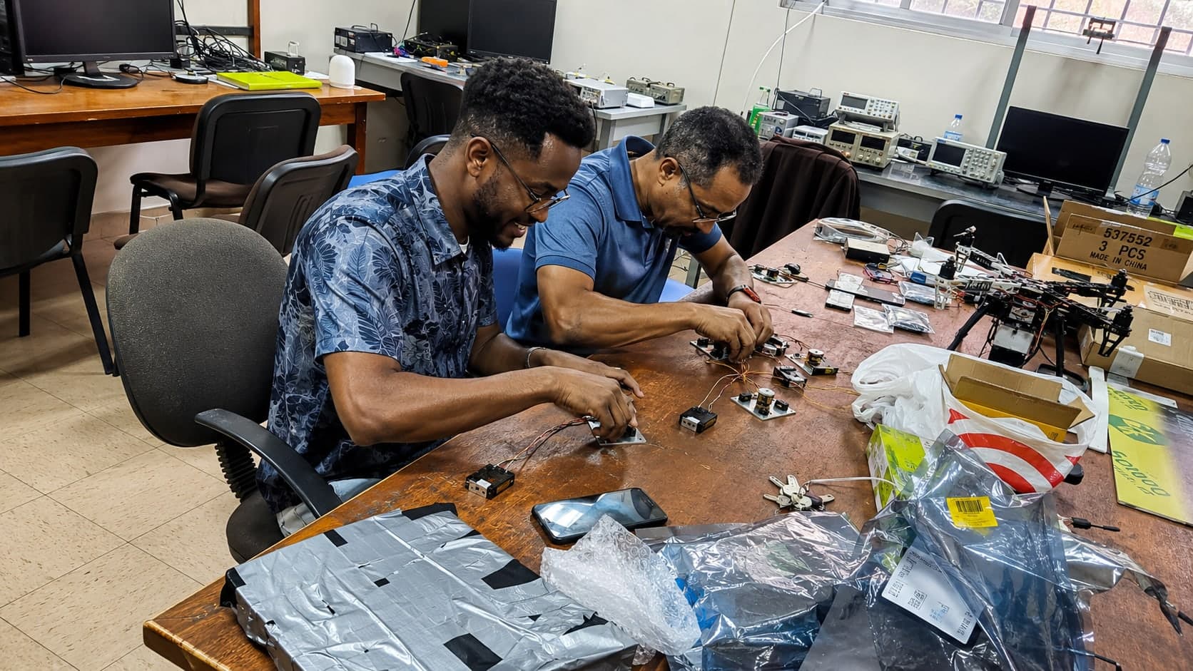 Two engineers at a university electronics bench wiring sensor modules by hand, with an oscilloscope, bench power supply and anti-static packaging around them.